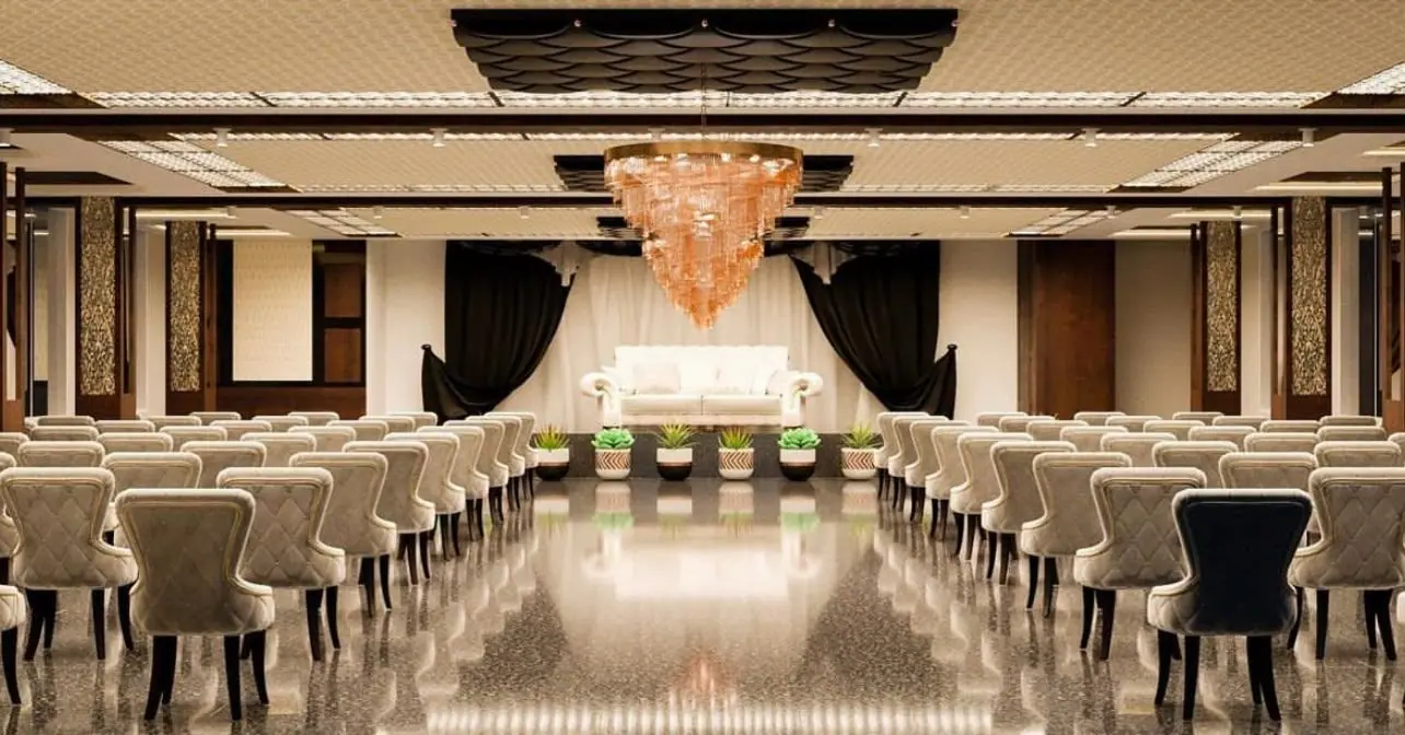 Banquet hall with tufted chairs either side of a centre aisle leading to a stage with a white sofa, drapes and a copper-toned crystal chandelier