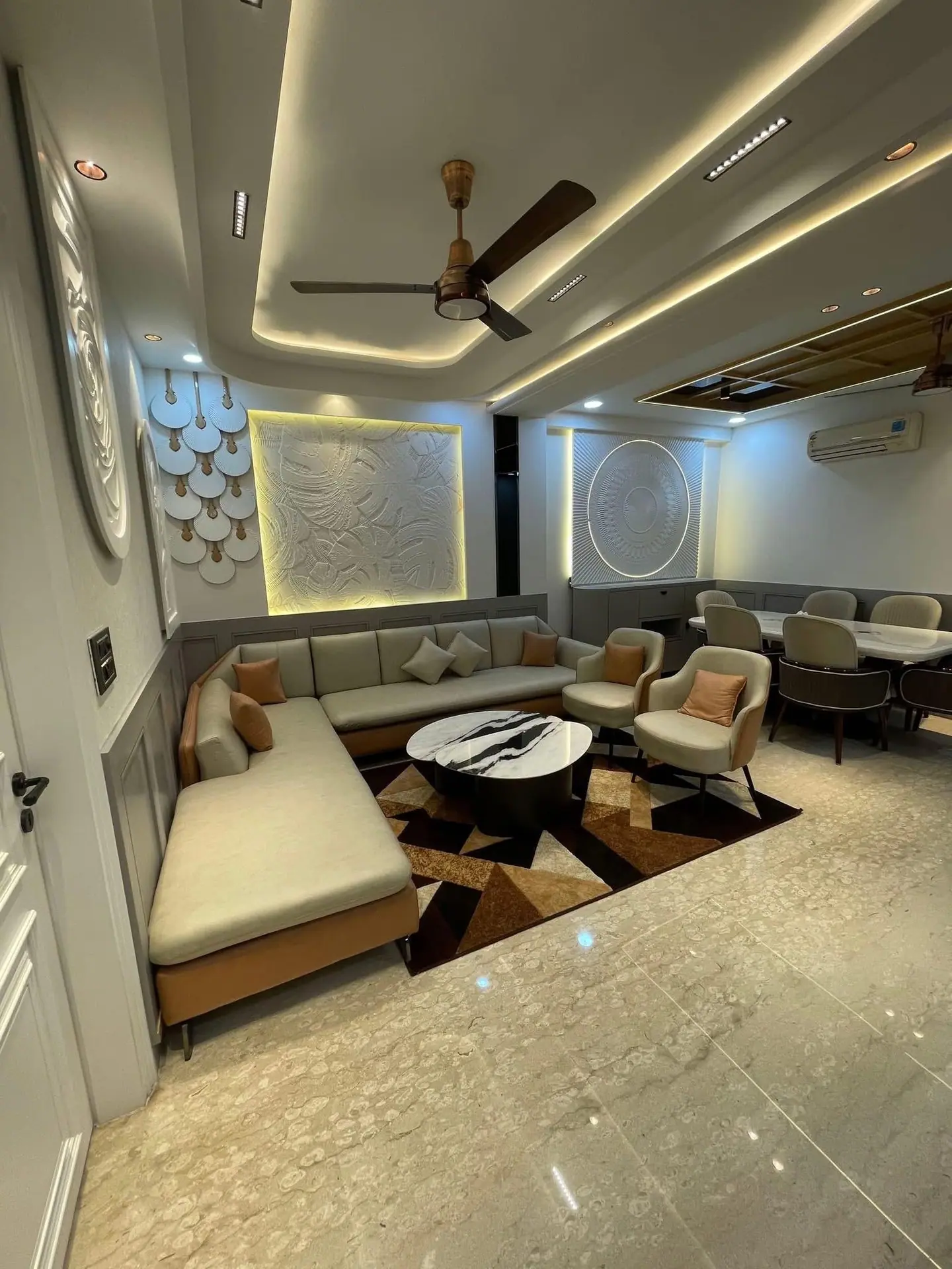 Living room with an L-shaped sofa, sculpted wall panels and a layered cove-lit ceiling, opening onto the dining area
