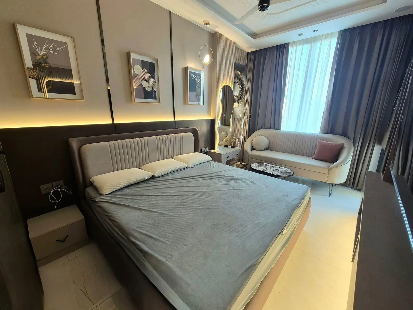 Bedroom with a channel-stitched bed, framed artwork above a lit wall ledge and a curved sofa by the window